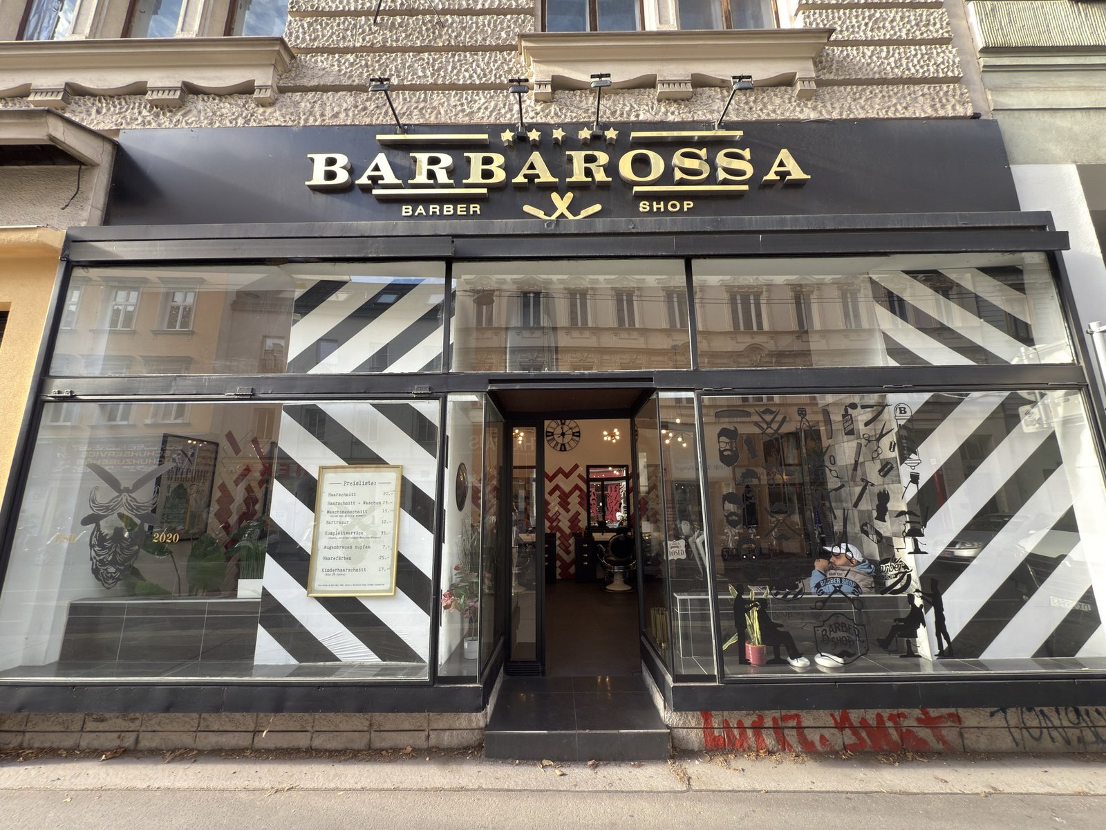 Fassade des Barbarossa Barber Shop mit goldenem Schriftzug auf schwarzer Tafel und gestreiften Schaufenstern