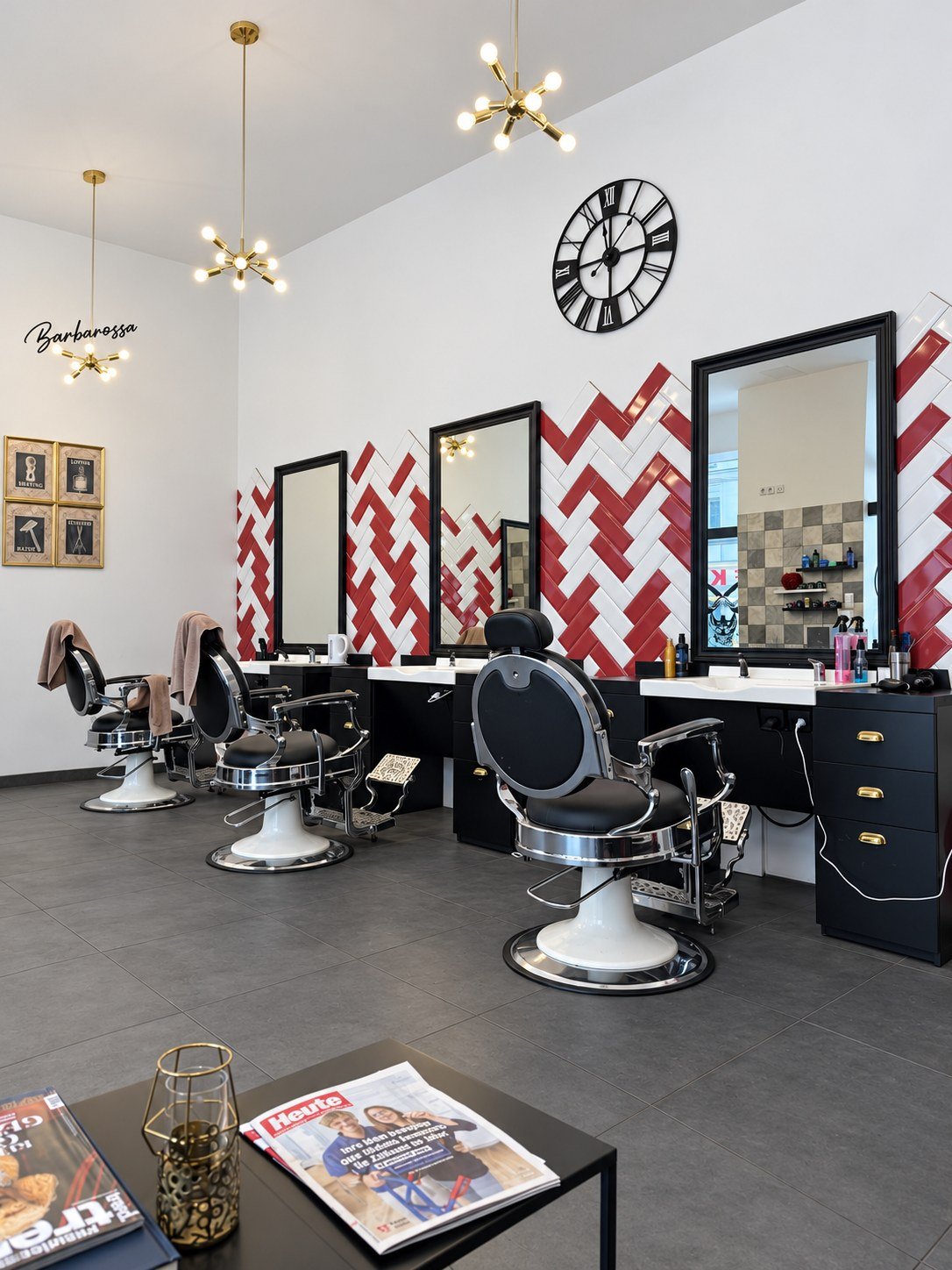 Blick in den Salon: schwarze Barbierstühle mit Chrom vor einer rot-weißen Fischgrätwand mit großen Spiegeln und Messing-Leuchten