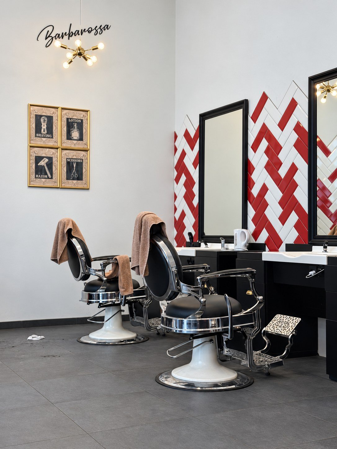 Zwei Barbierstühle mit Handtüchern vor der rot-weißen Fliesenwand, daneben gerahmte Barber-Poster und eine Messing-Leuchte