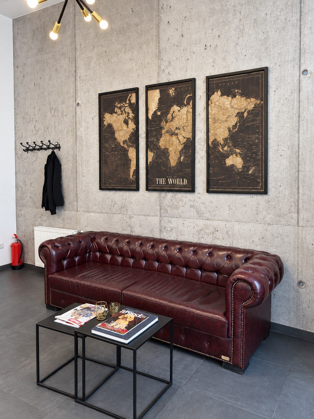 Wartebereich mit bordeauxrotem Chesterfield-Sofa vor einer Betonwand und drei gerahmten Weltkarten